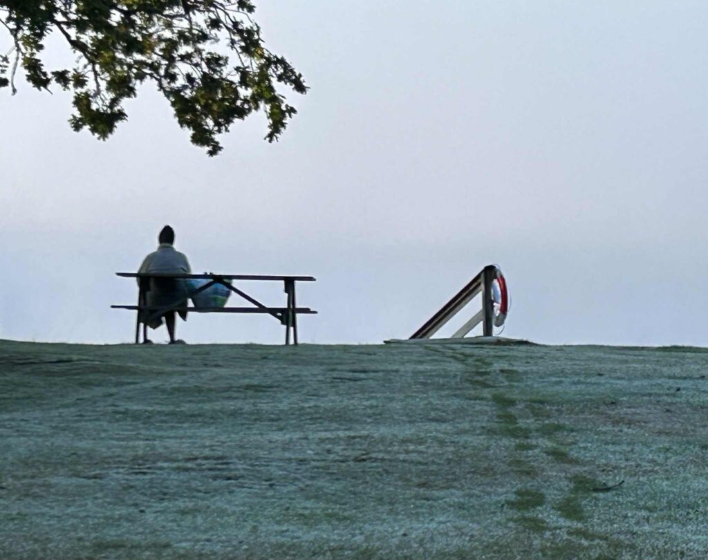 Morgonbad och tacksamhet – person insvept i morgonrock sitter på frostig bänk vid sjön en tidig höstmorgon, stillhet och dimma över vattnet.
