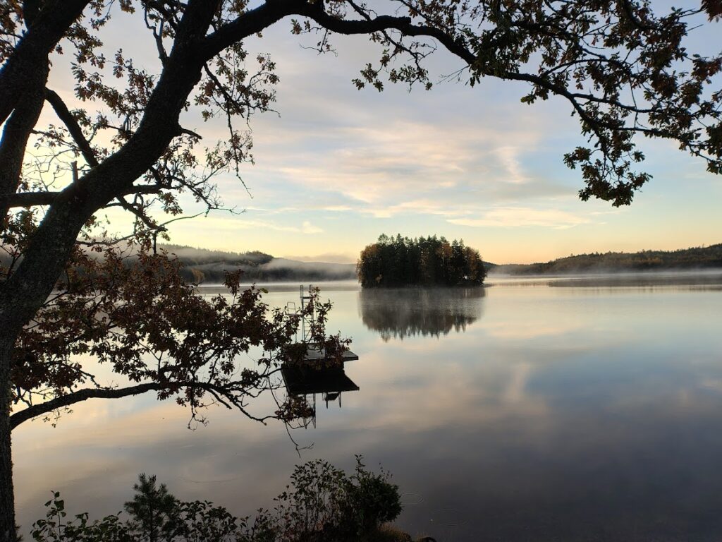 Morning mist over a mirror-still lake surrounded by autumn-colored trees and a small raft by the shore – a quiet moment reflecting why I write: presence, stillness, and life in motion.