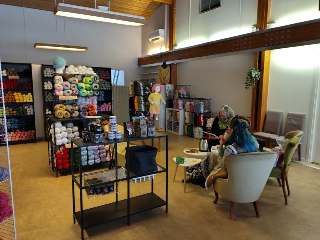 Två personer sitter i fåtöljer i en garnaffär, med garnhyllor runt omkring och en kaffekopp på bordet.