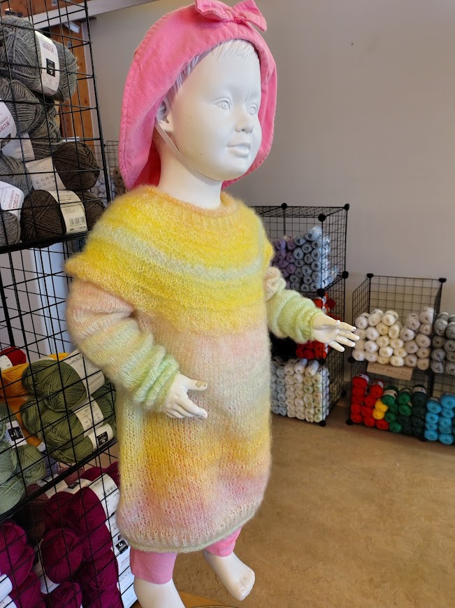 En docka i handstickad mohairklänning bland garnhyllor i garnaffären i byn.